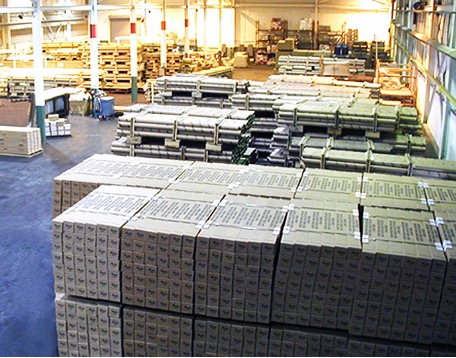 Neatly stacked boxes dominate the foreground in an indoor warehouse, while the background shows materials and equipment.
