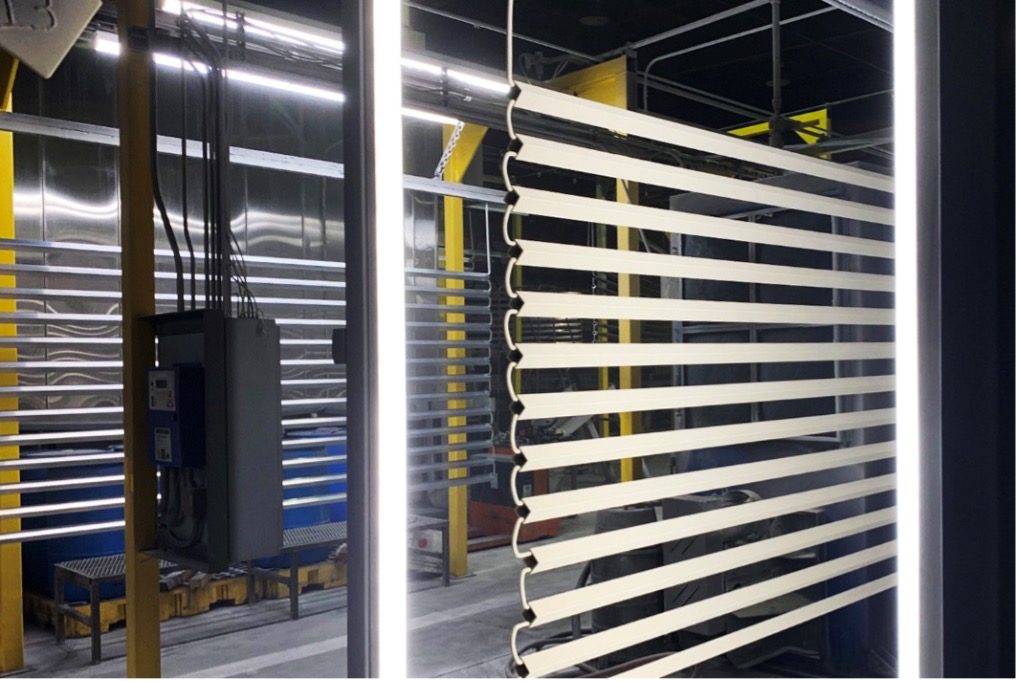 Wide image of the powder coating line with numerous long horizontal aluminum pieces hanging. One side shows aluminum extrusions without powder coating and the other side showing pieces that have just been sprayed in an eggshell white color.