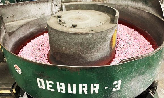 Green tumble deburr machine that reads “DEBURR – 3” on the side. It contains small pink colored ceramic media pellets.