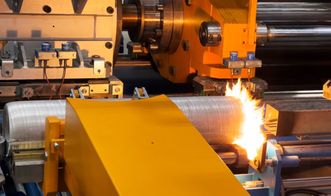 Image that shows a cylindrical long section on billet being prepared with heat to create soot to keep the billet from sticking when it goes into the extrusion press. Large orange section of metal holding the billet in place and flame being applied to and end of the billet piece.