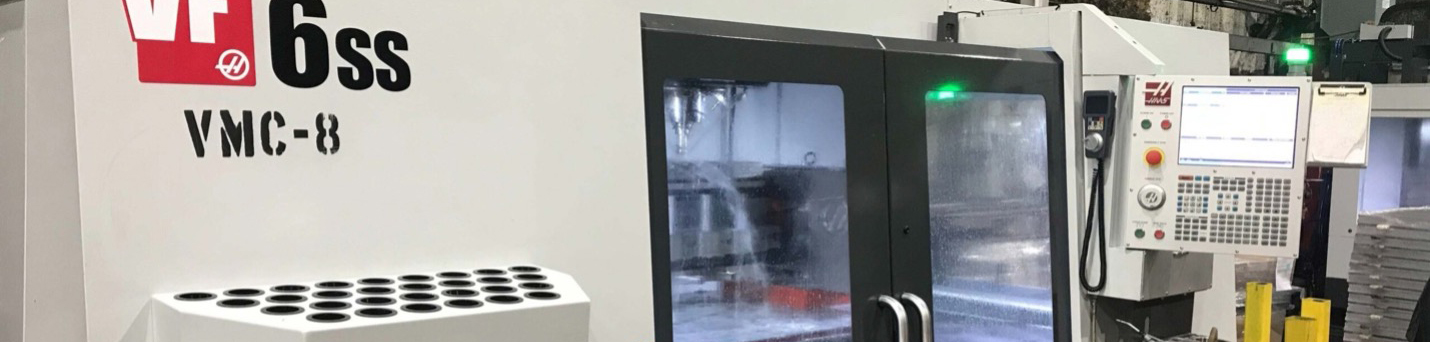 Wide angle of a CNC machine with a white metal case, glass front doors with black frames and a control computer screen for the equipment on the right side of the image.