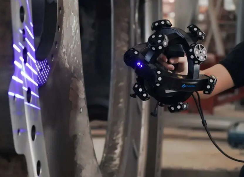 Image showing a TrackScan-P Tracking Scanner being held by a person on the right side of the image. Using it to scan an aluminum shape with purple laser lights being projected on the shape.